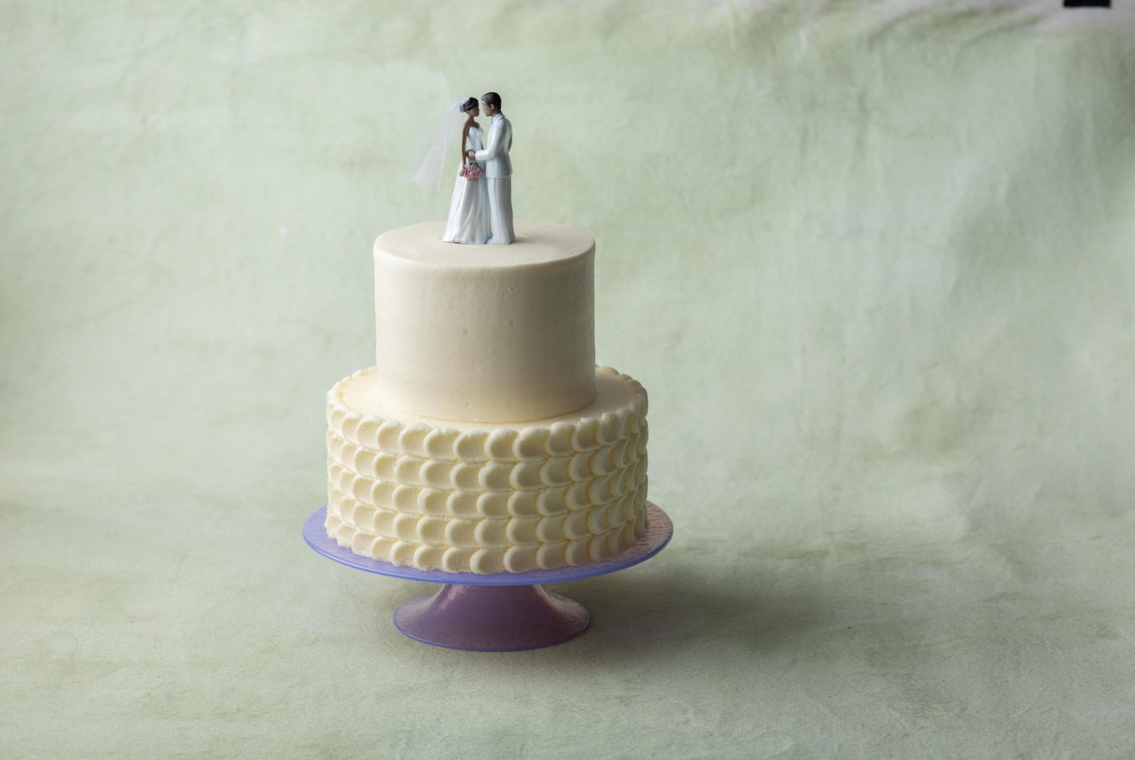A Magnolia Bakery two-tier classic wedding cake on a cake stand with traditional cake topper