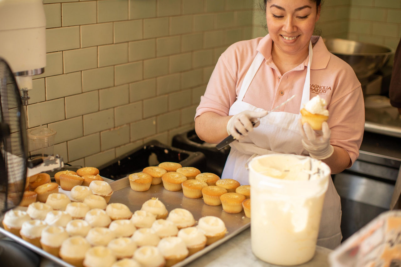 a tray of vanilla cupcakes being iced by a magnolia bakery employee